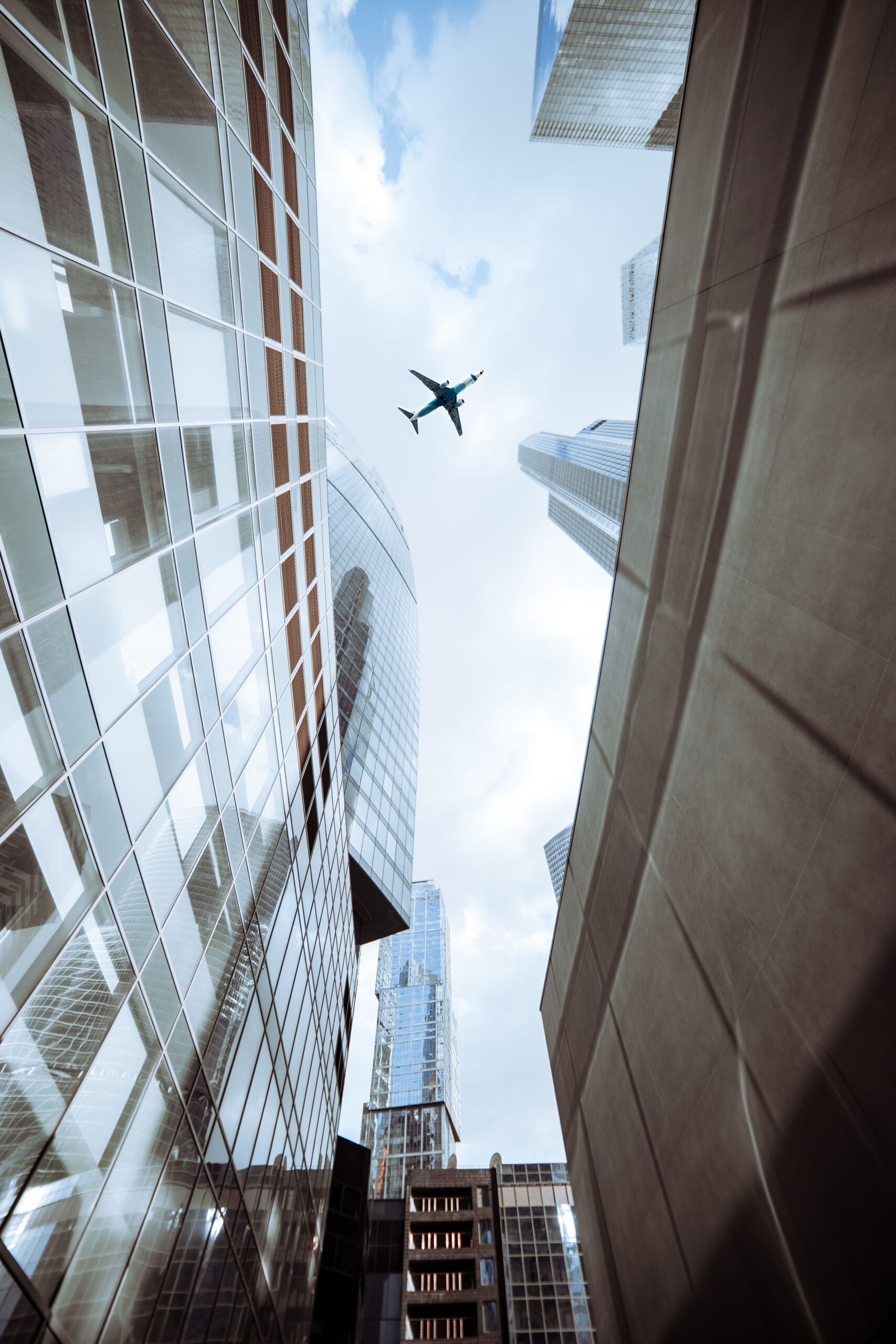 A dynamic perspective of an airplane soaring above city skyscrapers against a cloudy sky.